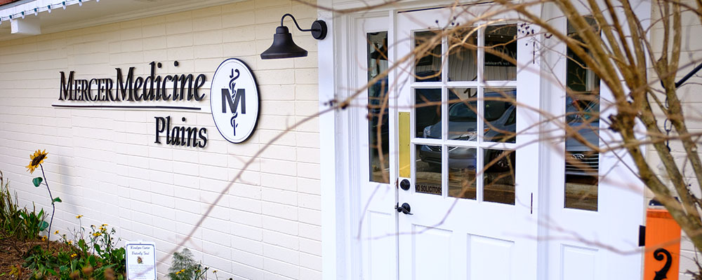 To the left of the door on a white brick building hangs a Mercer Medicine Plains sign as well as the Mercer Medicine logo.
